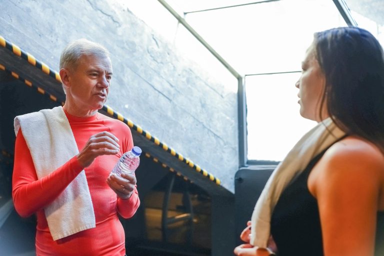 A senior man and a woman engaged in conversation in a gym, both in activewear holding water bottles.